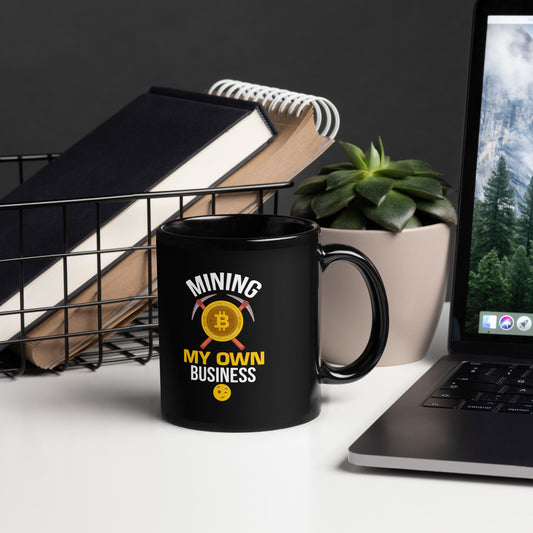 11 oz black glossy Bitcoin-themed coffee mug with “Mining My Own Business” design, placed on white desk beside books and laptop.